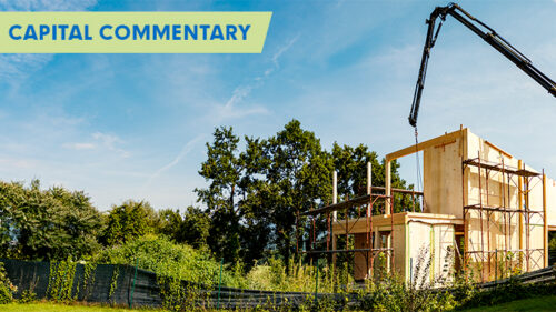 Panoramic view of a construction site with a wooden frame of a building under construction, a crane extending over the top, and a lush green landscape in the background under a clear blue sky. The text 'CAPITAL COMMENTARY' appears in bold letters in the upper left corner.