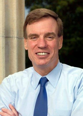 Senator Warner is facing the camera and smiling, showing this teeth. He is wearing a light blue button-up shirt with a dark blue tie.