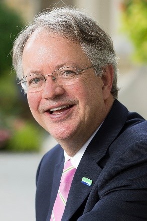 Tecklenburg is smiling directly at the camera with his body turned. He is wearing glasses, a black suit jacket, white dress shirt, and a pink tie.